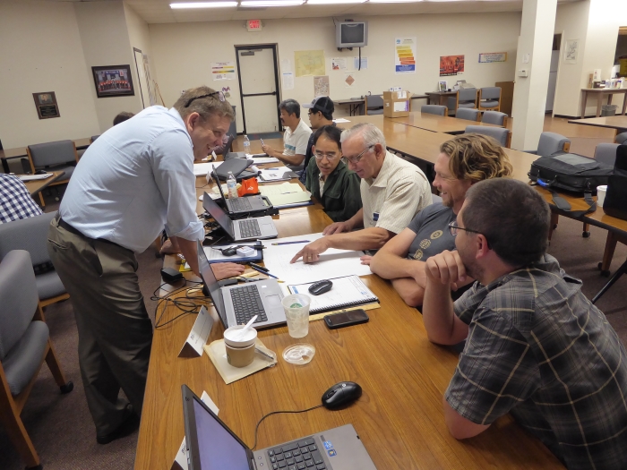 An instructor stands in front of a table across from several seated students. The student in the middle is pointing to a paper on the table.