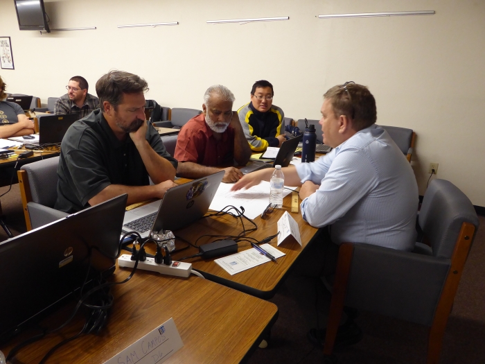 Three students face an instructor across a table. The instructor is pointing at papers on the table and talking.