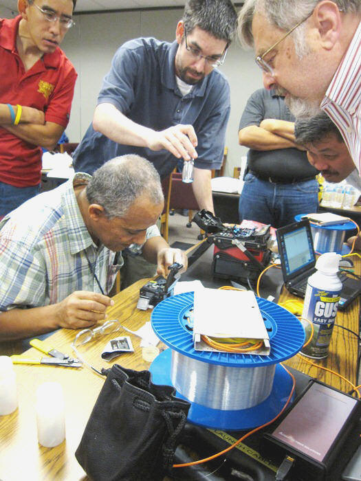 Berhanu Zergaw works carefully on a fusion splice while the instructor observes his work.  Other students gathered around to watch.  Clockwise from left:  Steven Gee, Michael Mullen, Dave Le, Eric Pearson