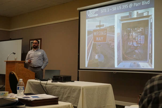 Man speaking next to a projector screen displaying a wrong way sign and the inside of a controller cabinet, slide text: Site 1 - SB US 395 @ Parr Blvd