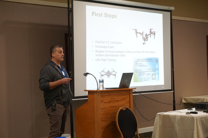 A man speaks behind a podium next to a projector screen displaying a picture of a drone and the words First Steps