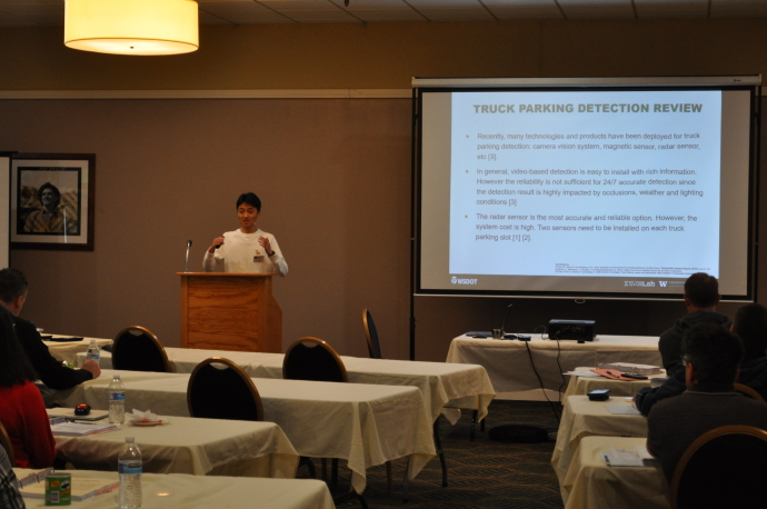 A speaker presents at a podium beside a screen showing Truck Parking Detection Review, audience watches