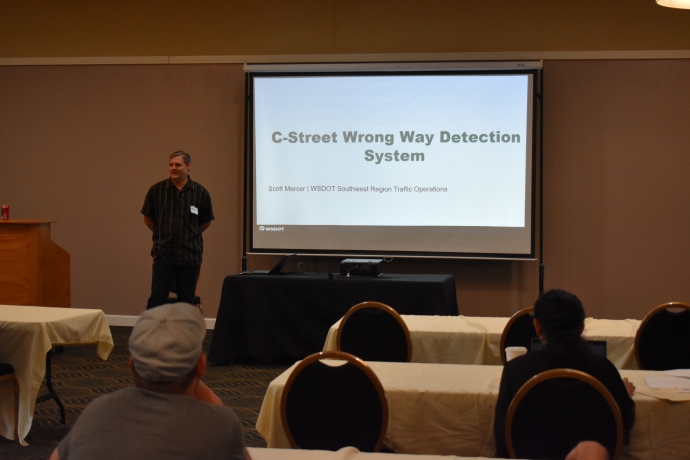 A man stands to the left of a projector screen speaking to an audience. The screen is titled 'C-Street Wrong Way Detection System.'