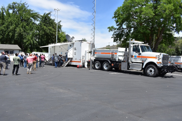 Caltrans satellite communications truck and trailer parked in a parking lot, antennas and dish deployed, a group of people looking at truck/trailer and entering trailer.