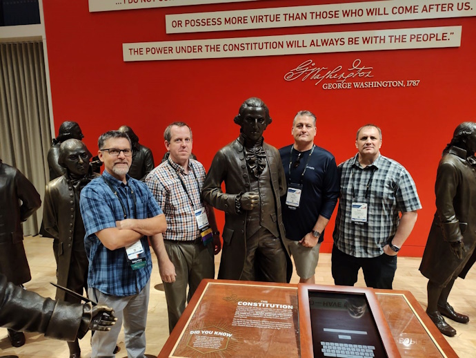 Several life-size statues with four men posing among them. In front is a display about the Constitution and on the wall behind is part of a quote from George Washington in 1787 – “or possess more virtue than those who will come after us. The power under the Constitution will always be with the people.”