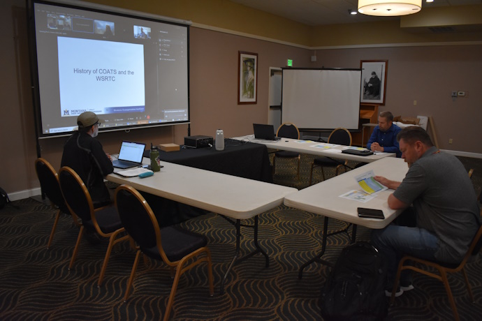 Three people seated at tables listen intently. A projector screen at the front shows online meeting participants and a slide with text: History of COATS and the WSRTC.