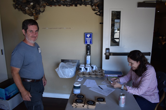 Left, a man standing in front of a table, a woman seated at the table on the right smiling and looking at a phone and a spreadsheet. Branded coasters and mugs on the table.