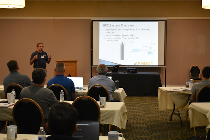 A man to the left of a projector screen gestures while speaking to an audience. The words “HFC System Overview” are on the screen with a picture of a cylinder tank.