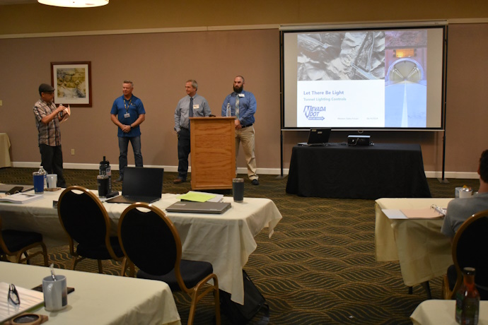 Four men stand beside a projector screen looking out to an audience. The slide on the screen has text “Let There Be Light, Nevada DOT”, a picture of a tunnel, and an overhead terrain view of a tunnel.