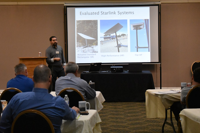 A man beside a projector screen speaking to an audience in front of him. On the screen, text = Evaluated Starlink Systems, three photos showing rectangular panel antennas on stands or mountings.