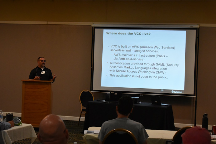 A man behind a podium beside a projector screen gestures at an audience in front of him. The screen has text = Where does the VCC live?