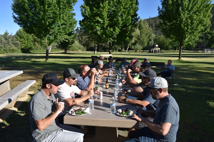 A long picnic table filled with people eating in a park with green grass and tall trees.