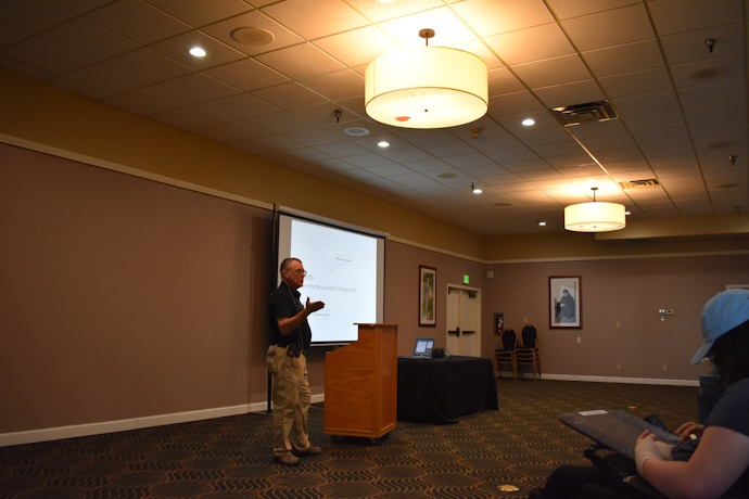 A man gestures while speaking beside a projector screen, laptop, and podium.