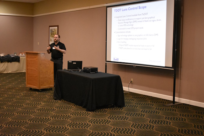 A man with a beard gestures with both hands while speaking behind a podium, projector screen to the right with text = TDOT Lane Control Scope.