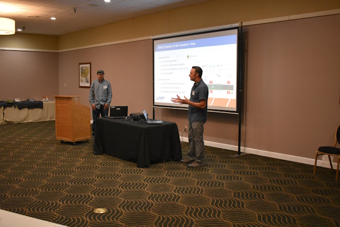 Two men stand on either side of a table with laptops, a projector screen in the background. The man on the right is gesturing while speaking.