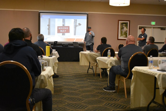 A man stands beside a projector screen with a “WRONG WAY” sign displayed. An audience looks towards the man.