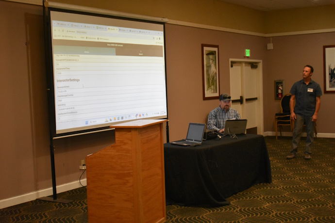 A man seated at a table operating a laptop with a projector screen showing a website application to his left; another man looks on to the right.