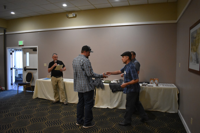 A man with a microphone stands in front of a table to the left. In the center, one person is handing another person an item.