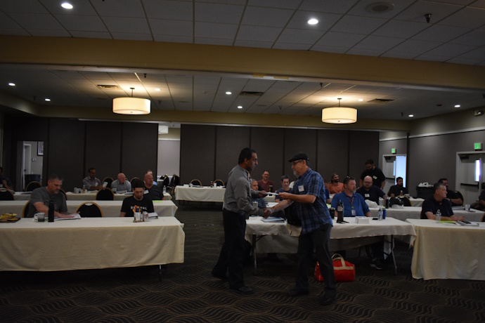 A man wearing a beret hands a man a piece of paper and they shake hands. An audience looks on and applauds.