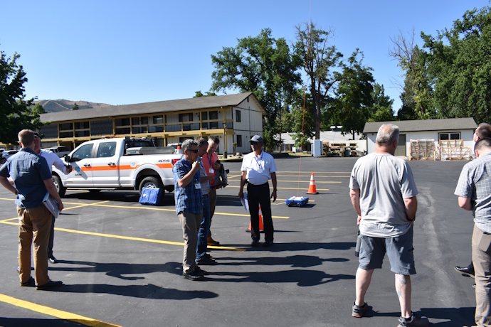 In a parking lot, six men gathered in three groups talking, one on the right looks skyward, one in the center with a clipboard and a white polo shirt is leading discussion, DOT pickup truck and Fotokite base station in the background.