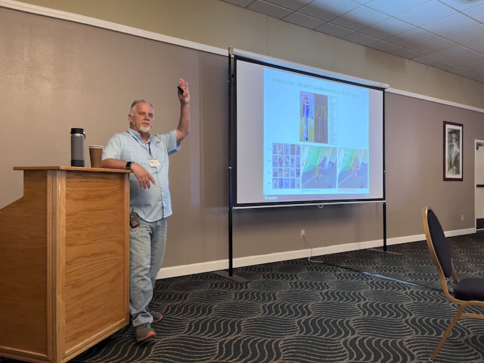 A man raises his left hand in the air, resting his right arm on a podium, while speaking beside a projector screen. He's dressed casually in a light blue shirt and denim pants. White or grey full goatee and mustache.