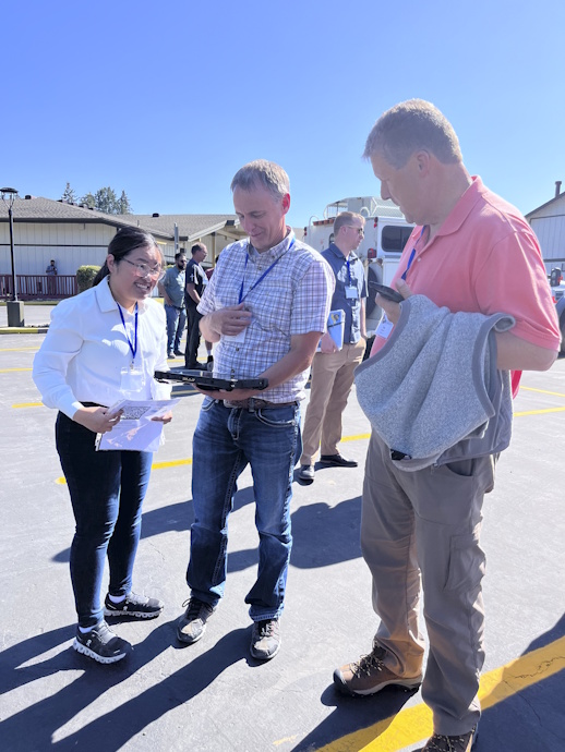 Outside in bright sunlight, three people stand around a tablet held by the man in the center. The woman on the left is responding to a question.