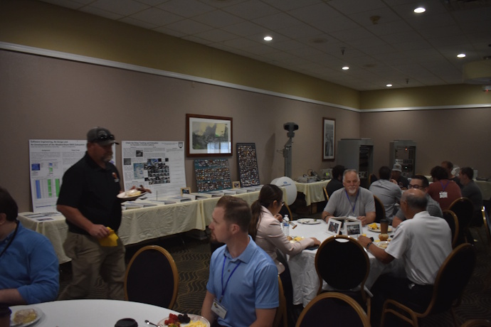 People gathered at round tables eating. Technical posters, photo collages, ITS equipment on tables in the back along the wall.