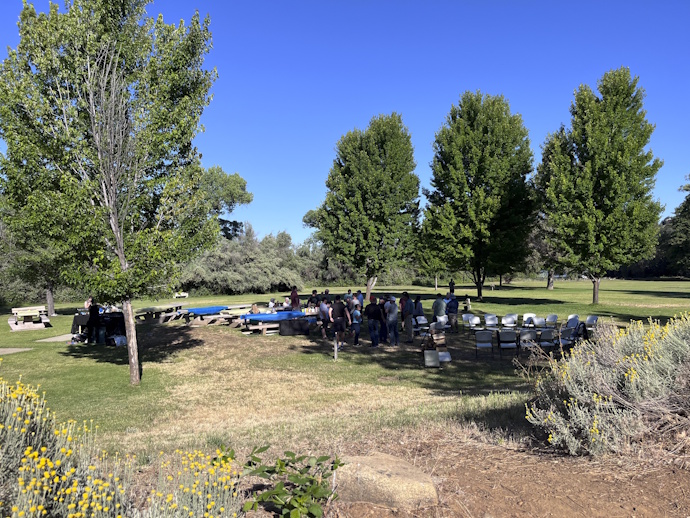 A group of people gathered at picnic tables and chairs in the shade of large trees, sunny with very blue sky, dryland vegetation in the foreground.