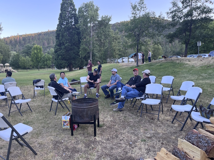 A group of people outside seated around a raised fire can; others standing in groups in the background.