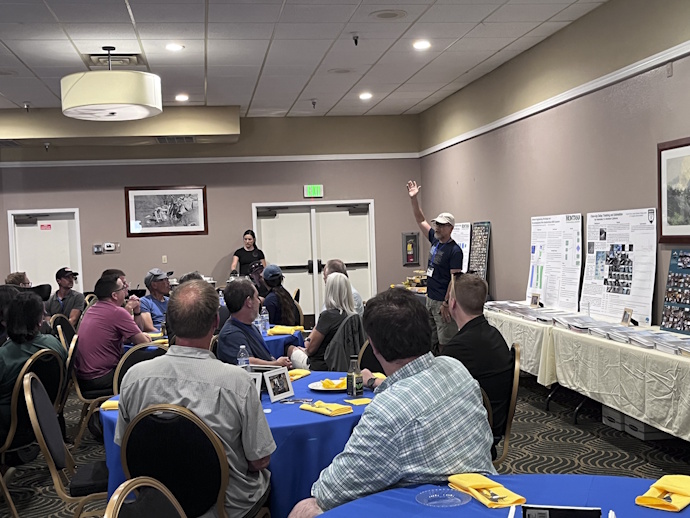 Groups of people at round tables covered by bright blue tablecloths with yellow napkins. They are looking to a man standing with his right hand in the air in welcome. Technical posters, 3-ring binders, and photo collages are along the wall on tables.