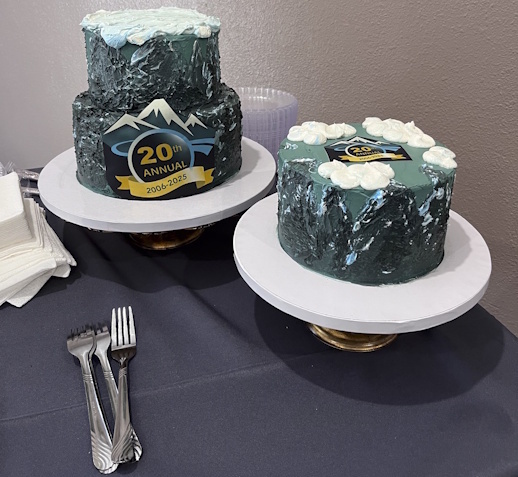 Two round cakes decorated in Forum colors on elevated turntables, the one on the left is double-decker and shows the 20th annual badge and mountain. Three forks are in front.