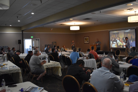 An audience looks to the front of a large room where three pen are standing and speaking beside a projector screen.